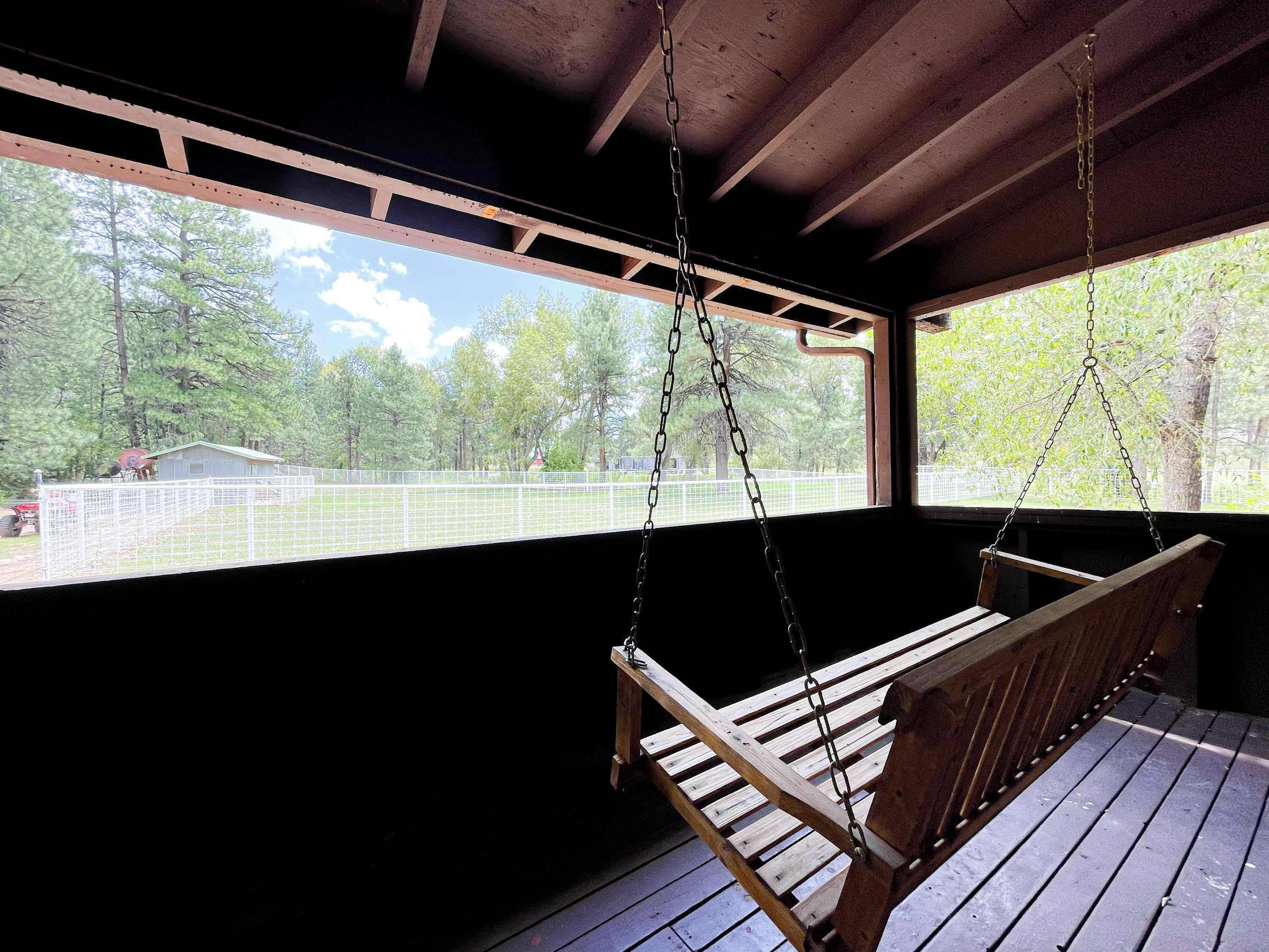 A wooden swing suspended from a ceiling in a covered outdoor area with a view of a fenced property and trees.