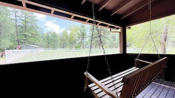 A wooden swing suspended from a ceiling in a covered outdoor area with a view of a fenced property and trees.