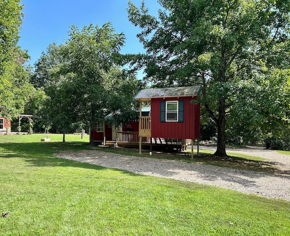 A red wooden cabin with a porch, surrounded by trees and a grassy area.