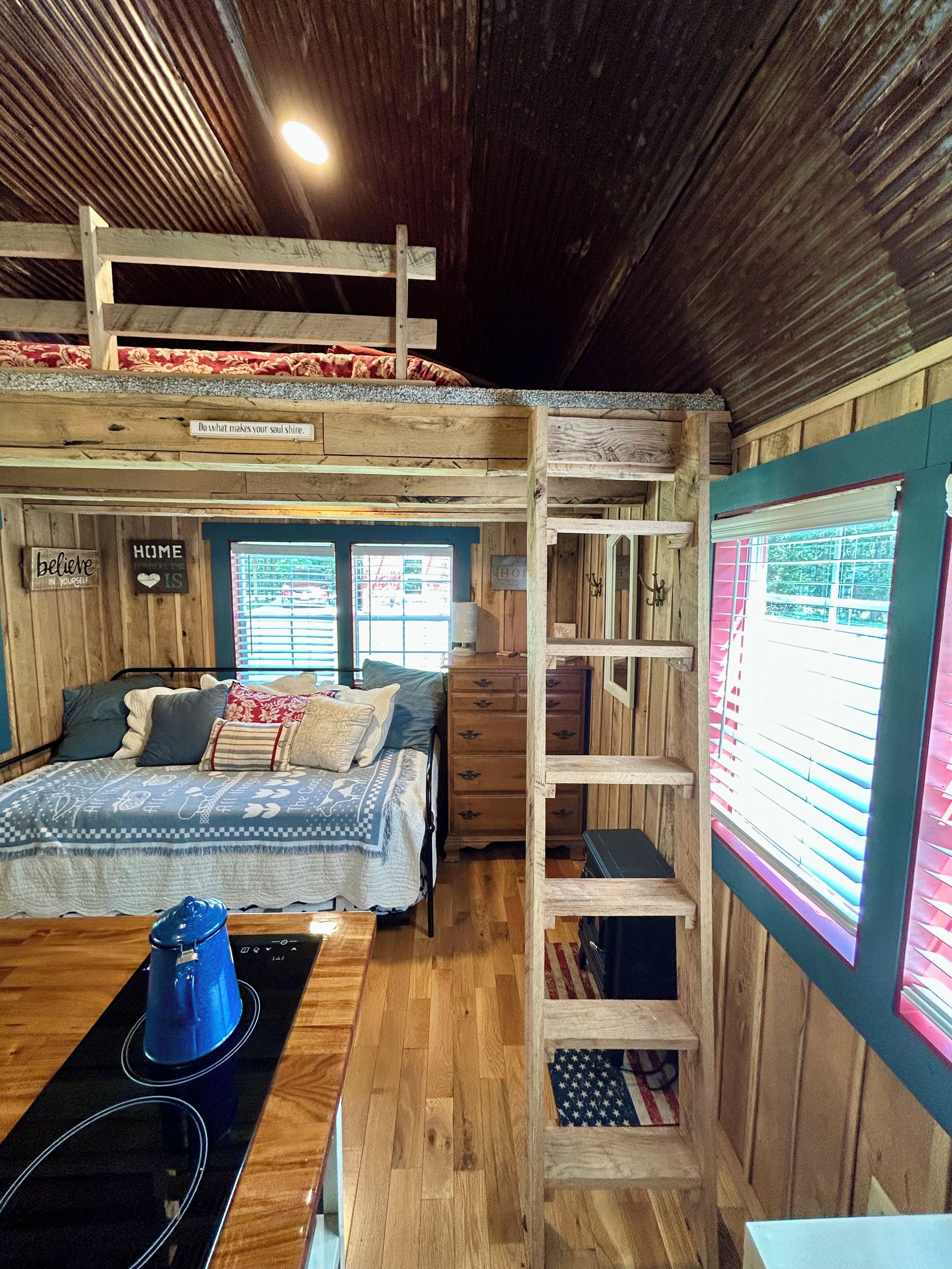 Een slaapkamer met stapelbed, houten plafond, bed met een bedekker met patroon, een tafeltje met een blauw voorwerp en een raam met jaloezieën.
