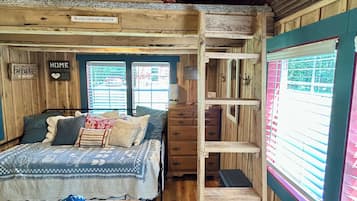 Een slaapkamer met stapelbed, houten plafond, bed met een bedekker met patroon, een tafeltje met een blauw voorwerp en een raam met jaloezieën.