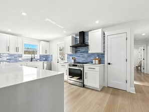 A modern kitchen with white cabinetry, a central island, a black range hood, and a blue-tiled backsplash. - New Design3 BDR Family Retreat (Jacksonville Beach)
