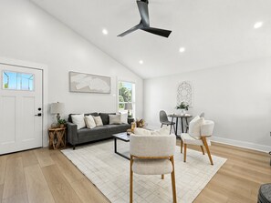 A modern living room with a grey sofa, two armchairs, a coffee table, and a dining table with chairs. There is a ceiling fan, a window with blinds, and a door. - New Design3 BDR Family Retreat (Jacksonville Beach)