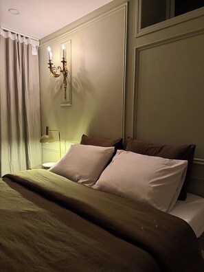 A neatly made bed with a green blanket and multiple pillows, a wall-mounted light fixture, and a curtain partially drawn to the side. - Stay at a remarkable English chateau style with a view & full optional services. (Calgary)