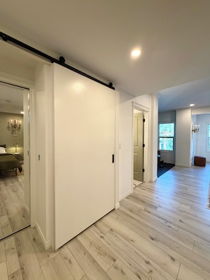 A spacious room with a large sliding door, wooden flooring, and a visible bed in the background.