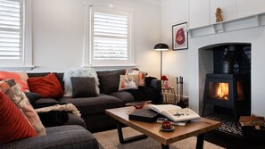 A cozy living room with a fireplace, a sofa with cushions, a coffee table with a book and mug, and a window with blinds. - Fontaine - Stunning 4 bedroom (Daylesford)