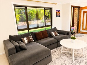 Ein modernes Wohnzimmer mit einer Eckcouch, einem runden Couchtisch und einem großen Fenster mit Blick auf einen Garten.