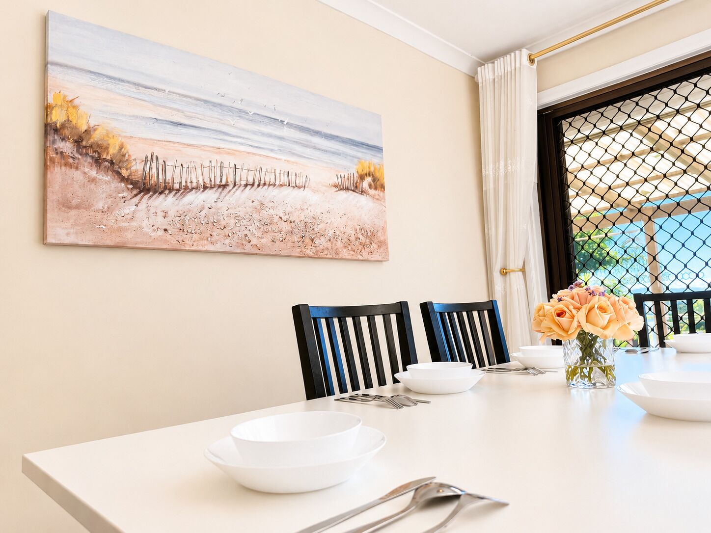 A dining area with a table set for two, chairs, a painting on the wall, and a window with curtains.