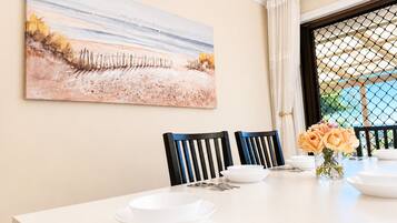 A dining area with a table set for two, chairs, a painting on the wall, and a window with curtains.