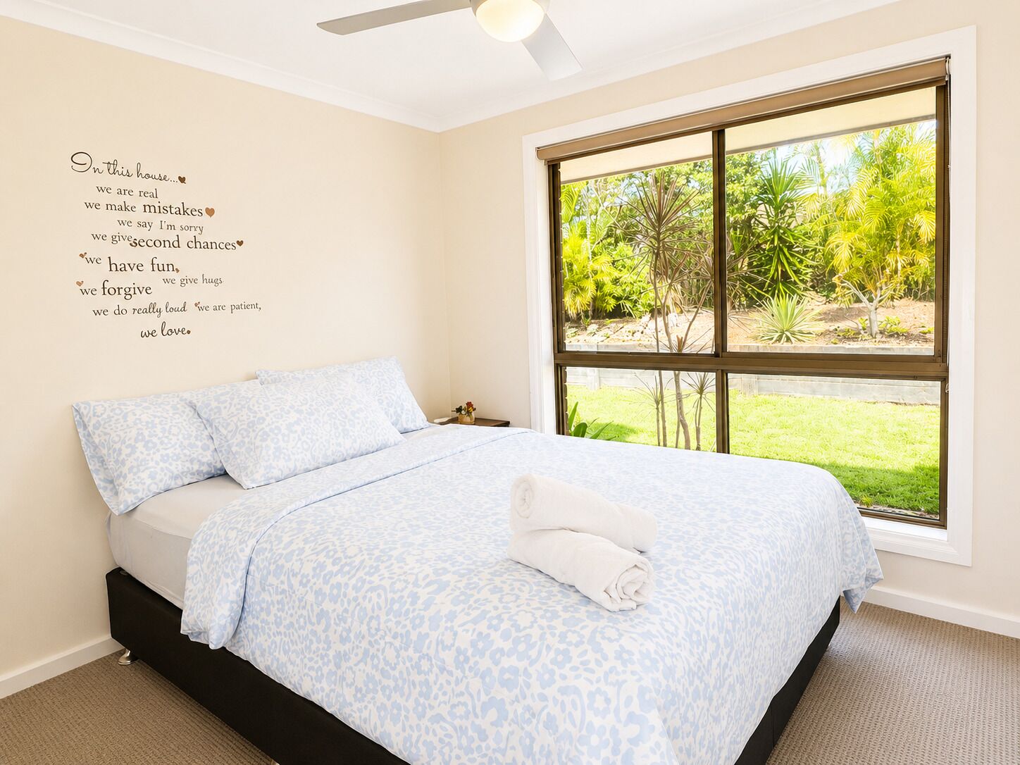 A bedroom with a bed, a ceiling fan, a window with a view of greenery, and a wall with text.