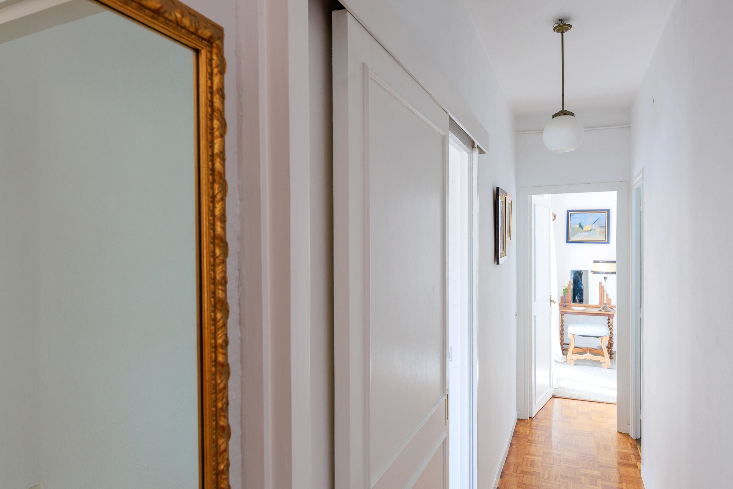 Un pasillo con una puerta a la izquierda, una pared blanca y un piso de madera.