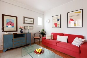 Ein Wohnzimmer mit einem roten Sofa, einem Glastisch mit Zitronen, einer blauen Kommode und verschiedenen gerahmten Kunstwerken an der Wand.