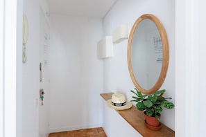 A hallway with a wooden floor, a white wall, a wooden-framed mirror, a wall-mounted lamp, a small shelf with a hat and a potted plant, and a door with a chain lock. - Pentahouse Banyoles, two terraces, fully furnished apartment can accommodate 2 guests (Barcelona)