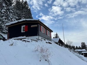 A two-story building with a dark facade and red shutters, surrounded by snow-covered trees and a clear sky. - Chalet Waldhüsli (CharmingStay), (Flumserberg Tannenboden), by Interhome (Flumserberg Tannenheim)