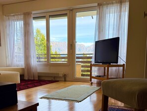 A room with a television on a wooden stand, a sofa, a red and white rug, and large windows with curtains. - Waldhaus Panorama Home (CharmingStay), (Flumserberg Tannenbodenalp), by Interhome (Flumserberg Tannenbodenalp)