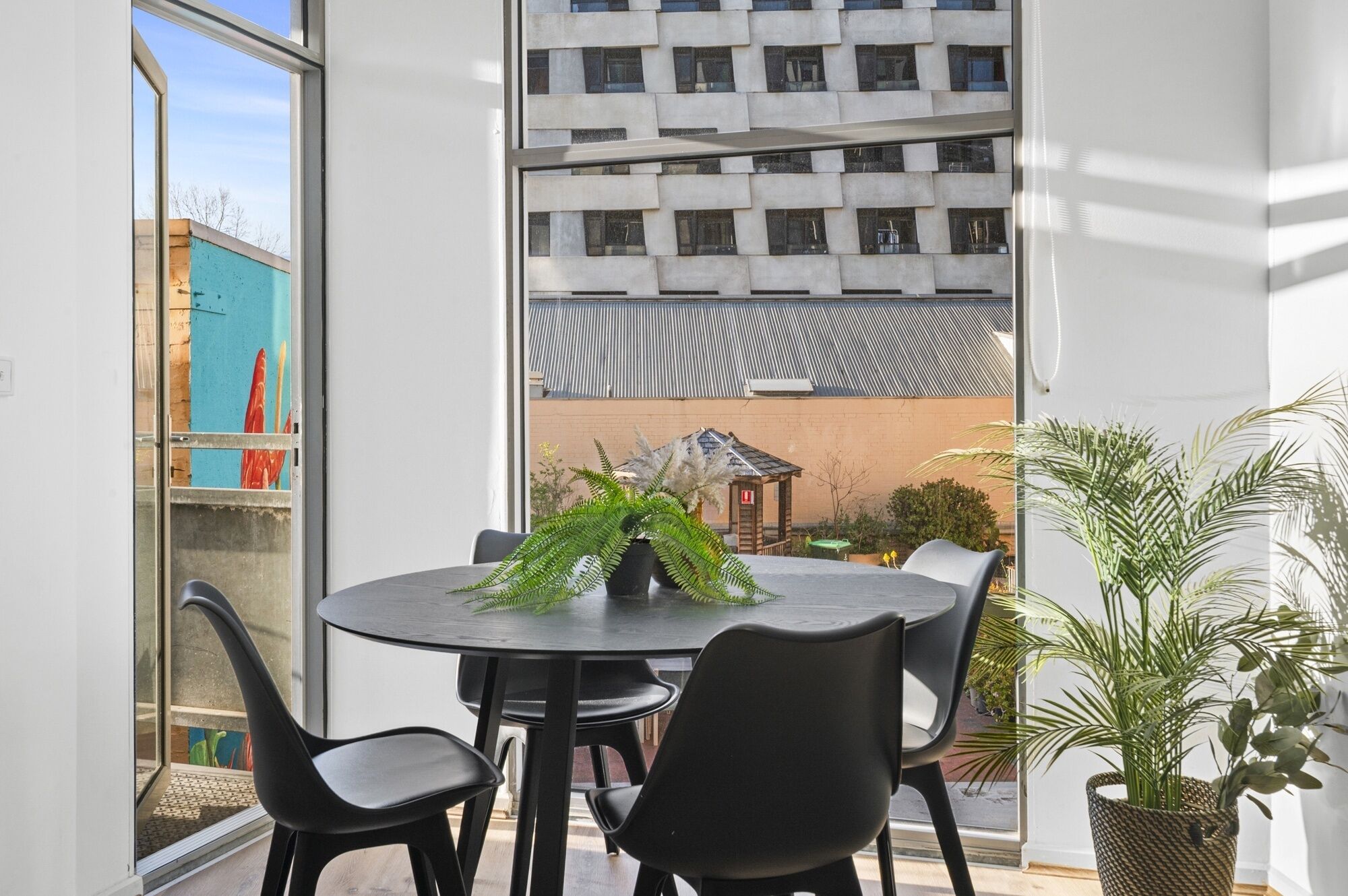 A modern room with a round table and four chairs, a potted plant, and a view of a building with a corrugated metal roof.