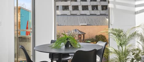 Quarto moderno com mesa redonda e quatro cadeiras, um vaso de planta e vista para um edifício com telhado de metal corrugado.