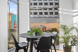A modern room with a round table and four chairs, a potted plant, and a view of a building with a corrugated metal roof. - Stylish 2-Bed Apartment Near Melbourne Central (Carlton)