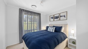 A bedroom with a bed, a nightstand, a lamp, and a window with curtains. - Nazza Daze in Narawallee (Narrawallee)