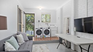 Ein kompakter Wohnbereich mit grauem Sofa, Flachbildfernseher, Waschmaschine und einer kleinen Küchenzeile.