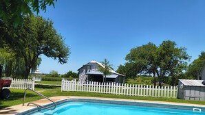 Piscina com cerca branca, um gazebo e um pequeno edifício ao fundo.