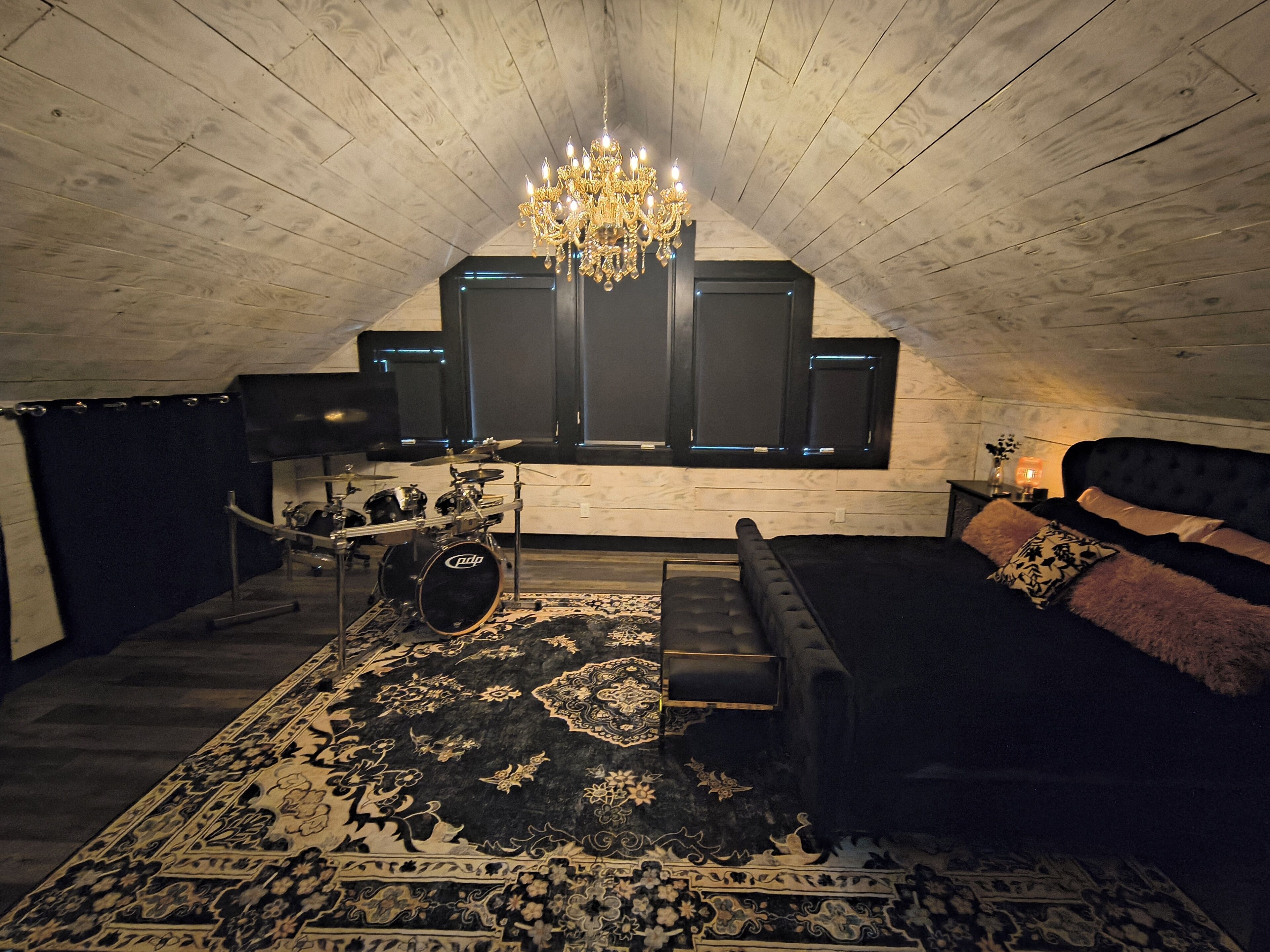 Ein Zimmer mit einer schwarzen Couch, einem gemusterten Teppich, einem Schlagzeug und einem Chandelier.