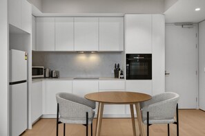 Eine moderne Küche mit weißen Schränken, einem schwarzen Ofen, einer Mikrowelle und einer Kaffeemaschine. Ein runder Holztisch wird von zwei grauen Polsterstühlen ergänzt.