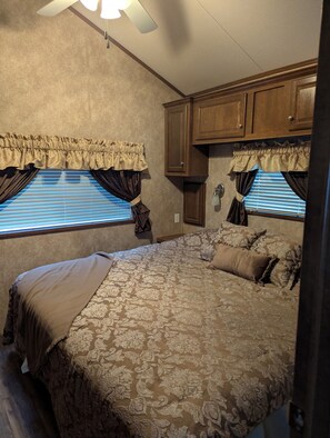 A bedroom with a large bed, two windows with curtains, a ceiling fan, and wooden cabinets.