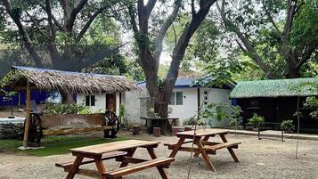 A rustic outdoor setting with wooden picnic tables, a thatched-roof structure, and a small building with a blue roof.