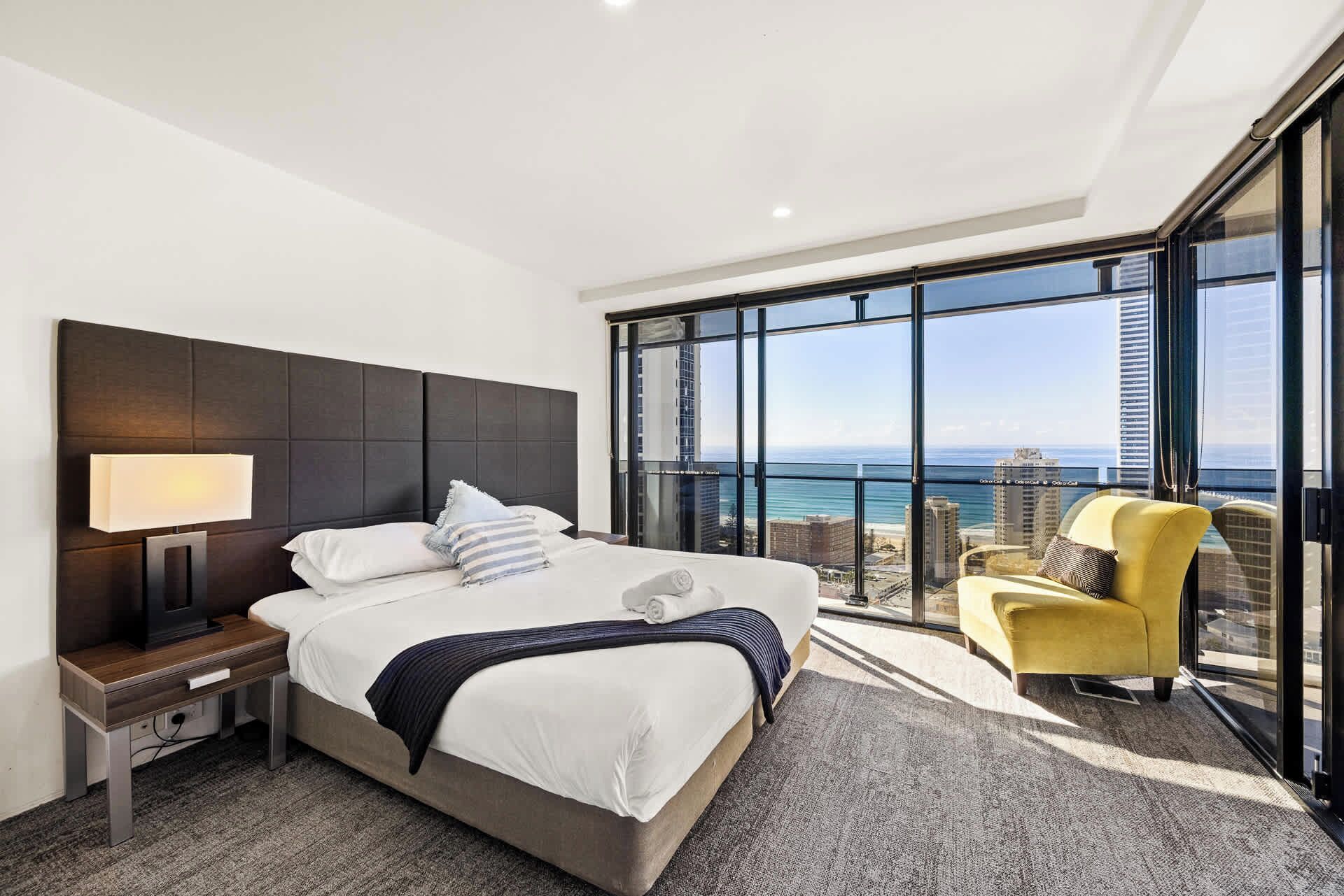 Ein modernes Hotelzimmer mit einem großen Bett, einem Nachttisch, einer Lampe, einem Sessel und einem Balkon mit Blick auf das Meer und die Stadt.