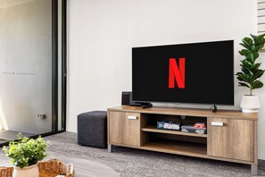 Un salon moderne avec un téléviseur à écran plat affichant le logo de Netflix, un meuble de rangement en bois sur lequel est posé un jeu d’échecs, et une plante en pot.