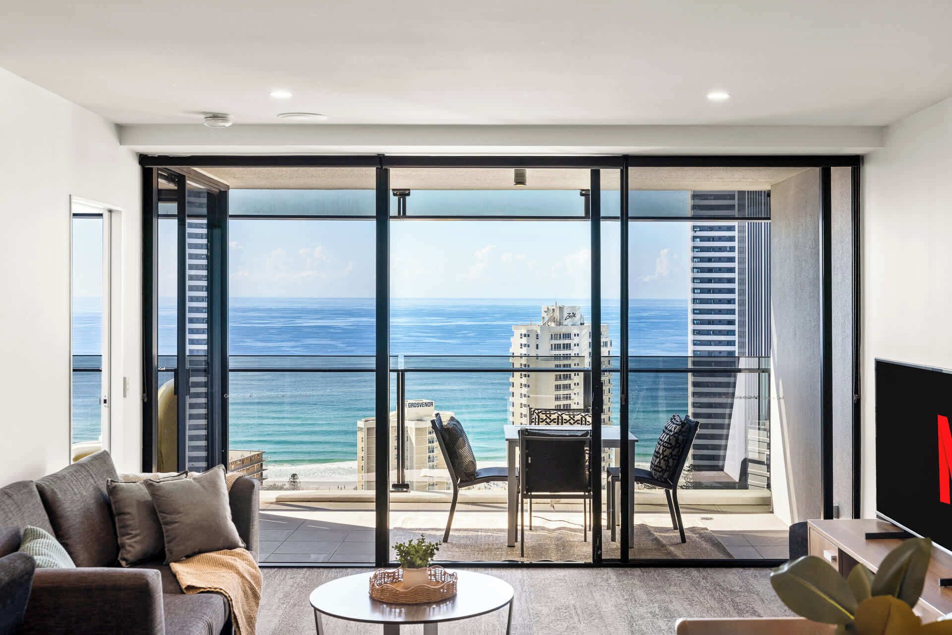 Ein modernes Wohnzimmer mit einer großen Schiebeglasdoor zum Balkon mit Blick auf das Meer und ein Gebäude.