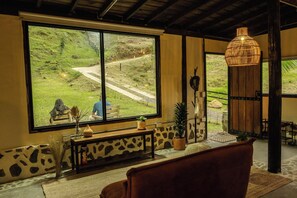 A room with a large window overlooking a rural landscape, a wooden bench, a table with a vase and a plant, and a hanging light fixture.
