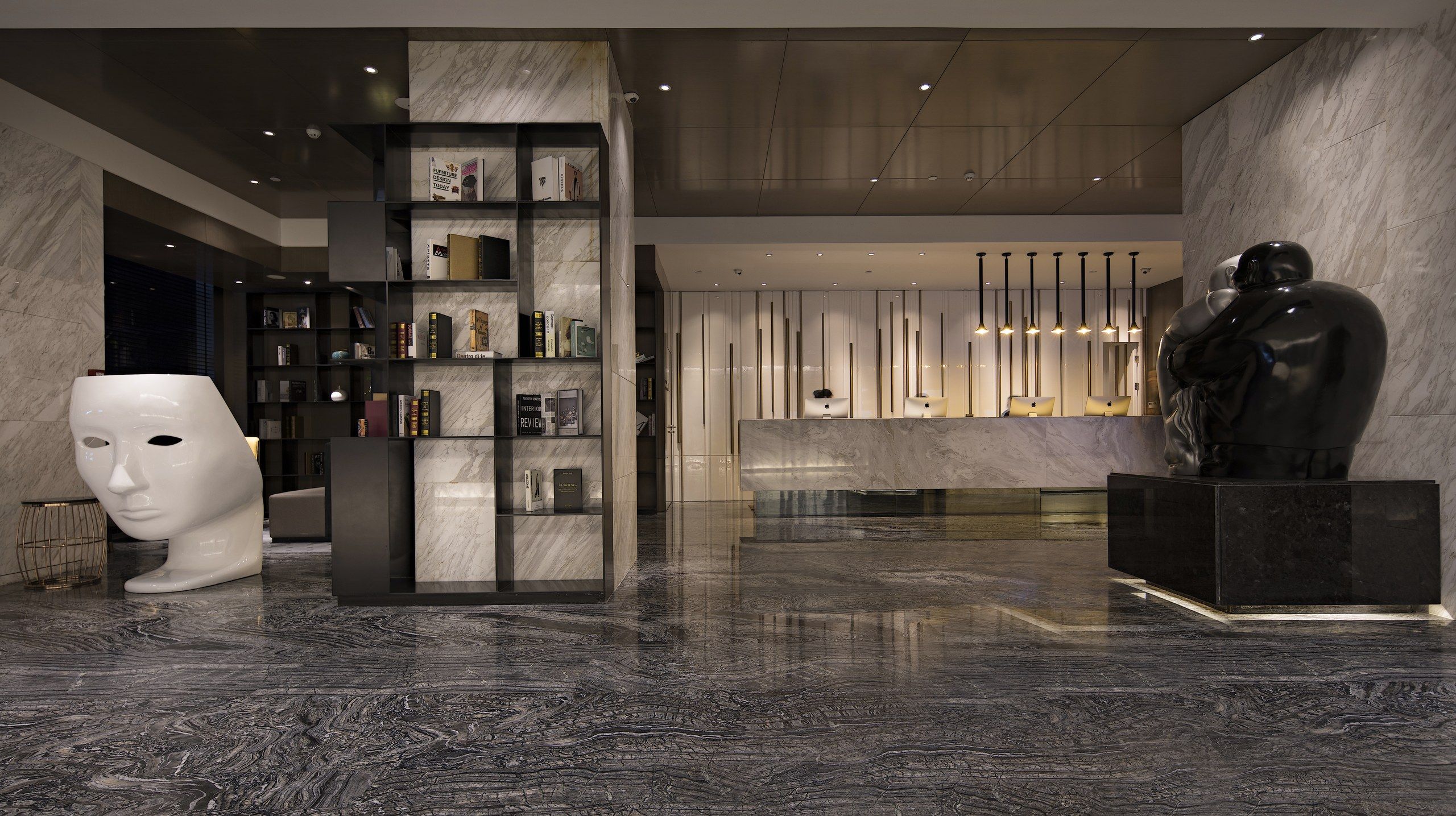 Een moderne lobby met een marmeren vloer, een grote boekenkast, een borstbeeld en een receptiebalie met verticale verlichtingsarmaturen.