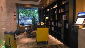Un hall de hotel con mostrador de recepción, una estantería, un área de descanso con sillas amarillas y un amplio ventanal con vista a árboles.