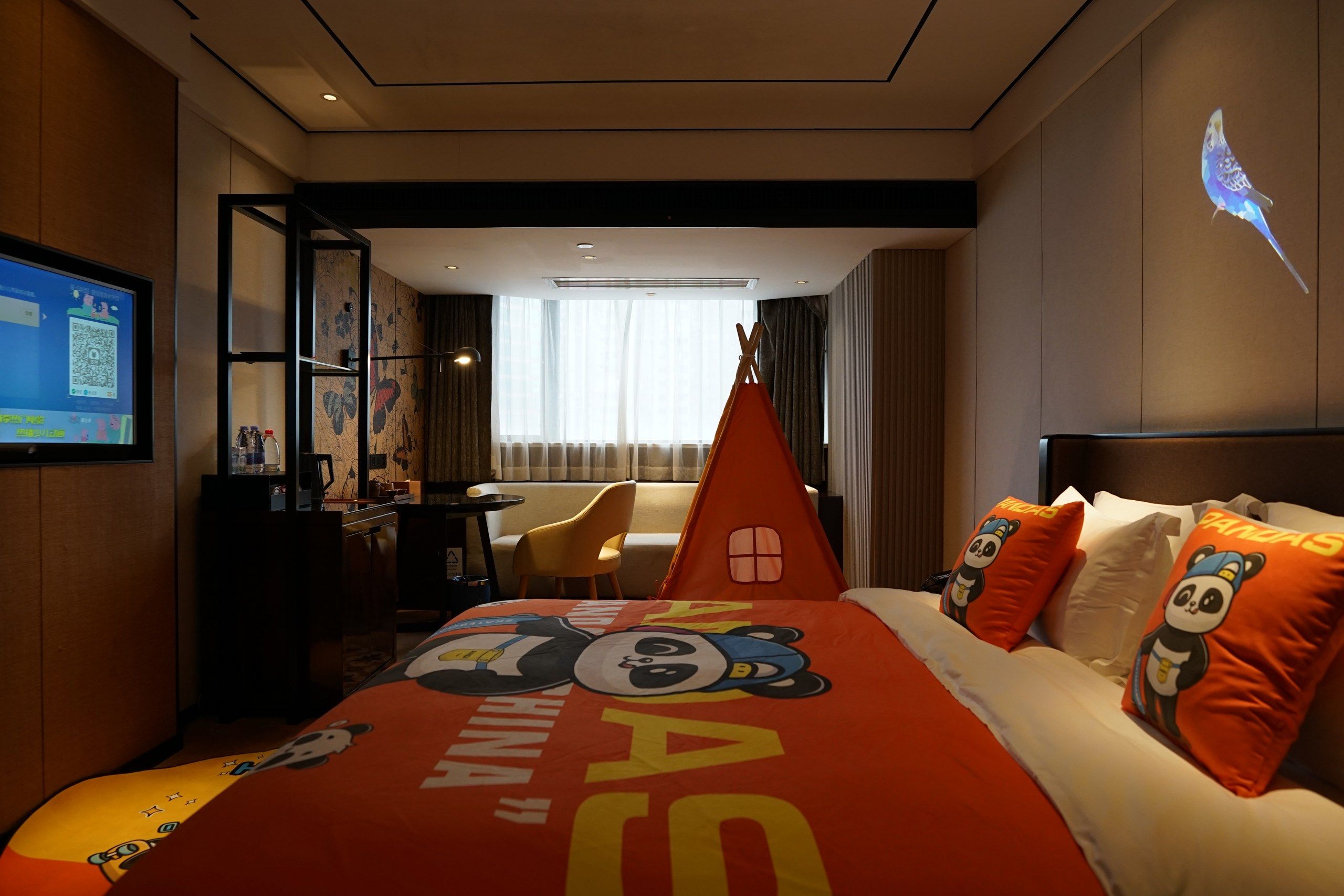 Une chambre d’hôtel avec un lit agrémenté d’une couvre-lit sur le thème des pandas, un bureau avec un ordinateur, une chaise et une grande fenêtre avec des rideaux.