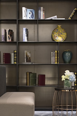 Uma estante com vários livros e objetos decorativos, um vaso com flores e uma cadeira.