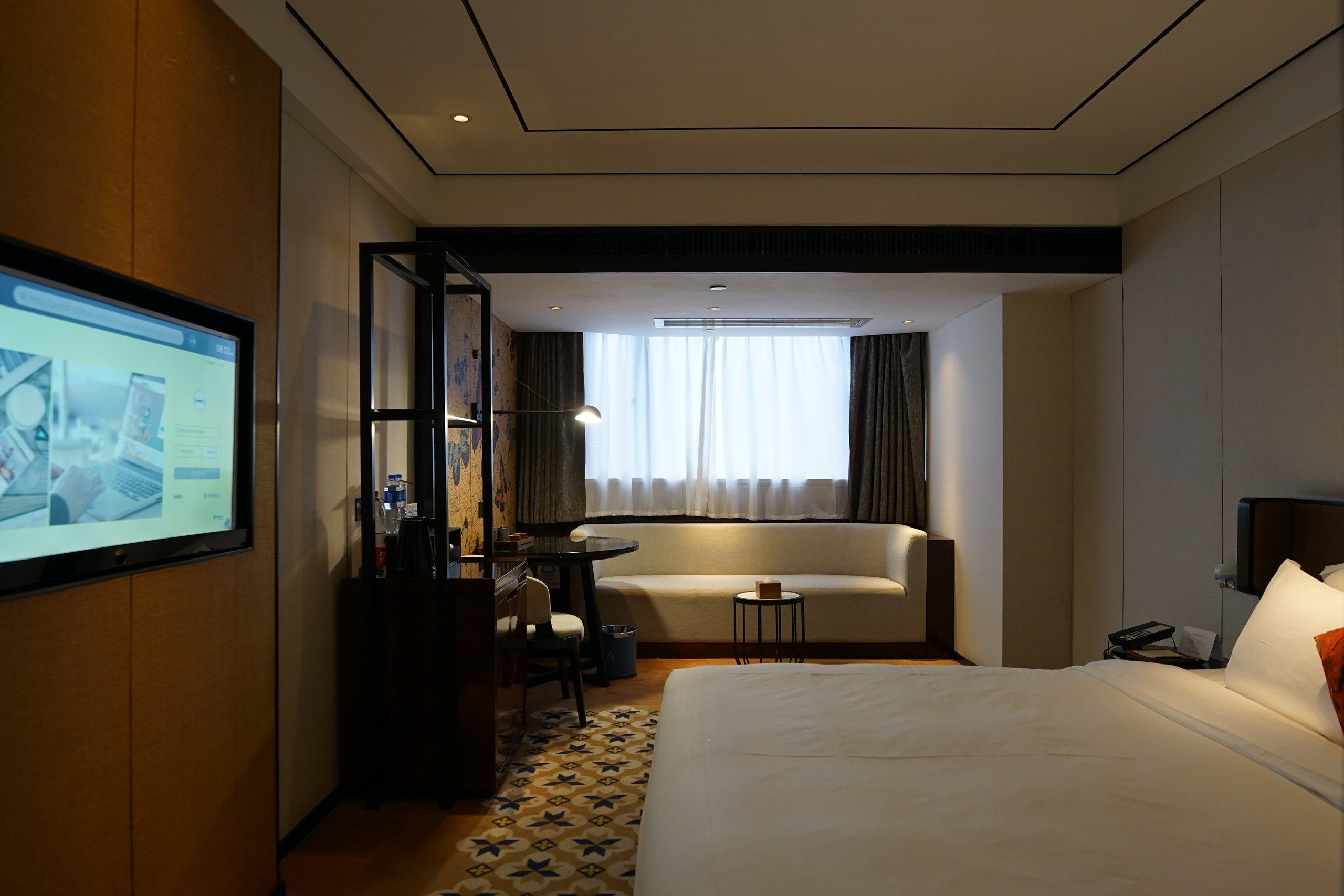 Una habitación de hotel con una cama, un escritorio con una silla, una mesita, un sofá y un televisor instalado en la pared.