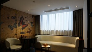 Una habitación con un sofá, dos sillas, una mesita, una pared con diseño floral, una ventana con cortinas y un respiradero en el techo.