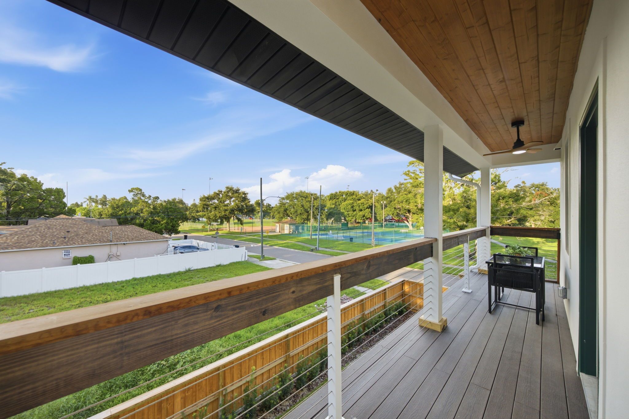 A covered outdoor deck with a view of a tennis court and a grassy area.
