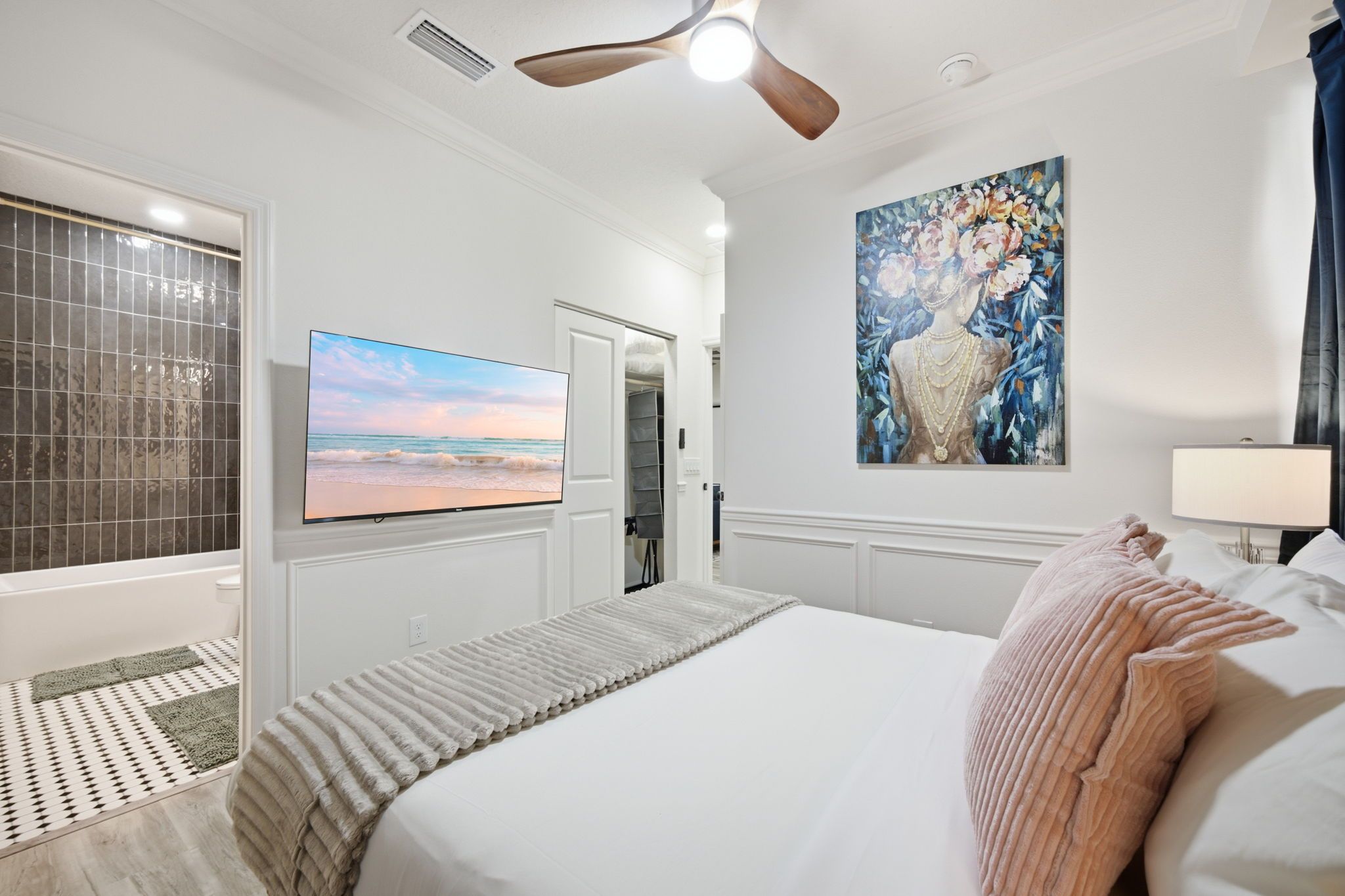 A hotel room with a large bed, a painting, a ceiling fan, and a view of the beach.