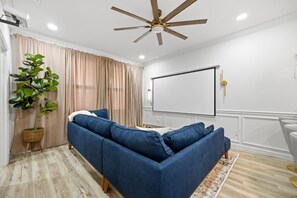 Ein Wohnzimmer mit Ecksofa, Deckenventilator, Projektionsleinwand und einer großen Topfpflanze.