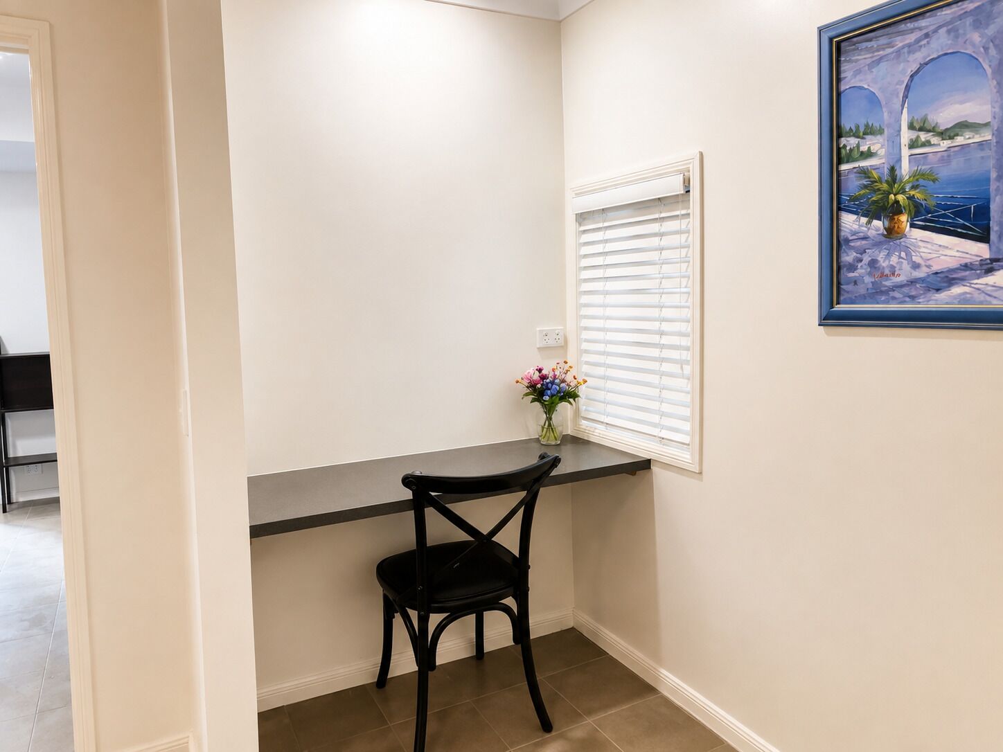 A room with a black chair, a small table, a window with blinds, and a framed painting on the wall.