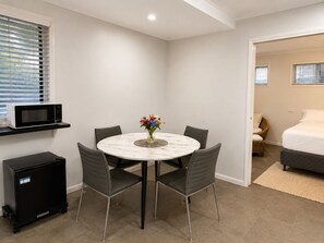 A compact room with a round dining table, four chairs, a microwave on a shelf, a small black cabinet, and a bed with a white mattress and pillows. - 1Bed 1Bath Ground floor West End (West End)