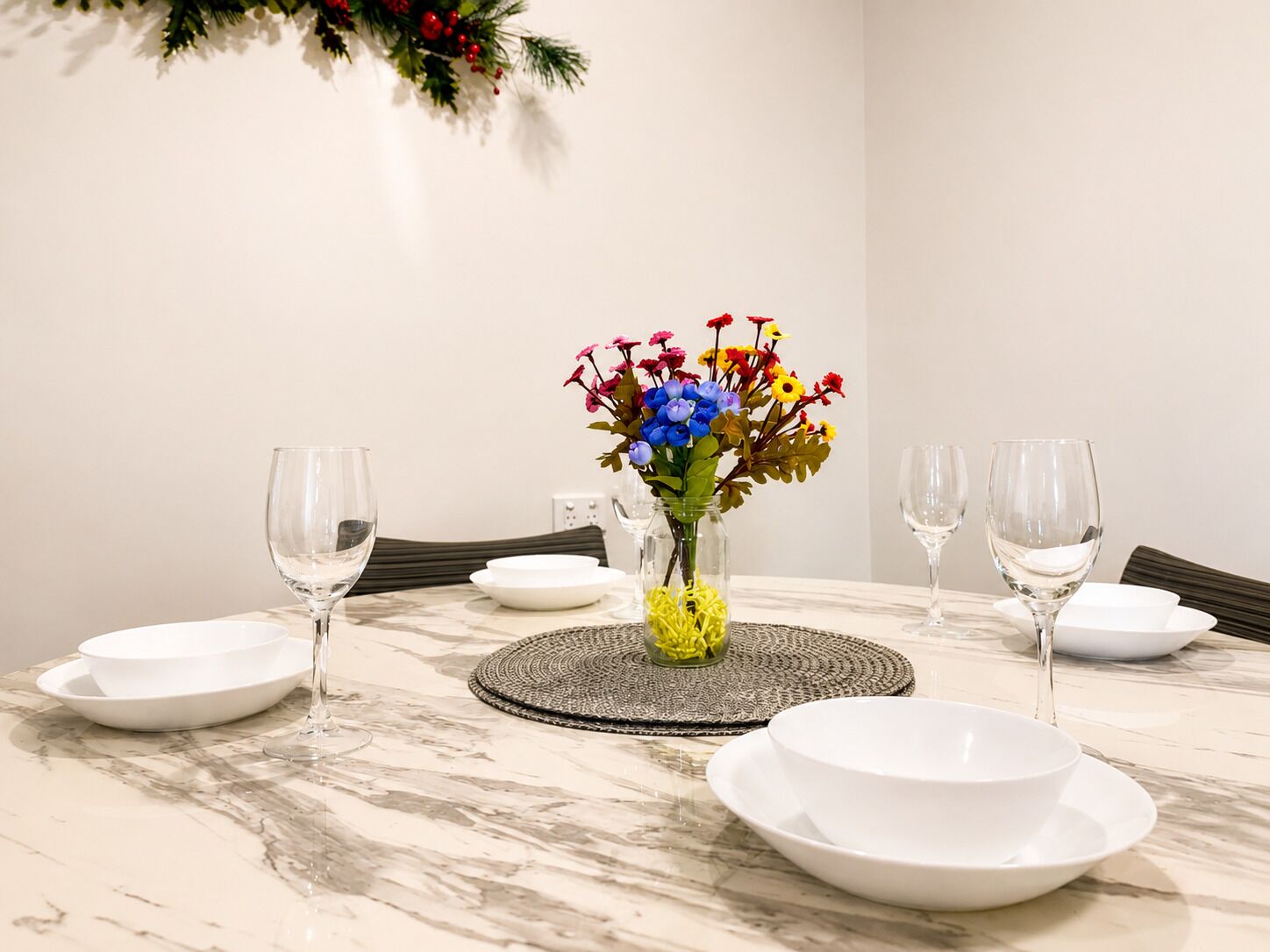 A dining table with white plates, clear wine glasses, and a vase with colorful flowers.