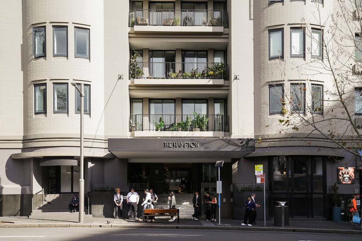 Ein mehrstöckiges Hotel mit einer geschwungenen Fassade, Balkonen mit Bepflanzung und einem Schild mit der Aufschrift 'THE HAMPTON'.
