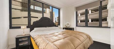 Un dormitorio con una cama grande, dos mesitas de noche, una mesilla auxiliar y una ventana con vistas a un edificio.