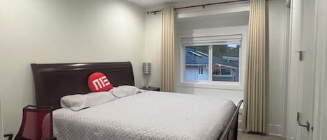 A hotel room with a bed, a nightstand, a red chair, a window with curtains, and a view of the outside.