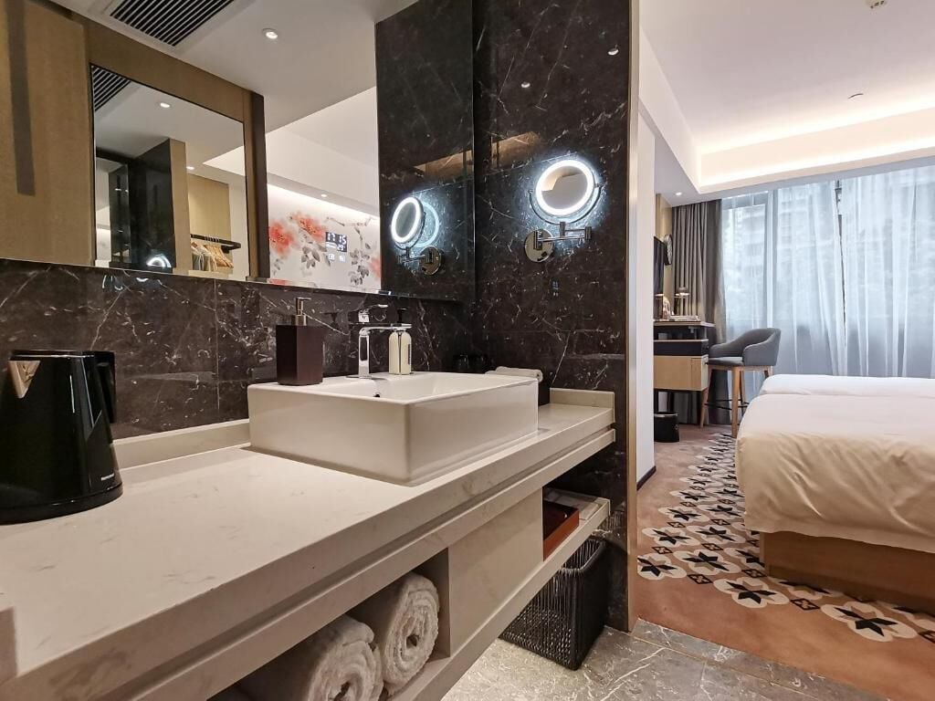 A modern hotel bathroom with a large mirror, two sinks, a coffee machine, and a towel rack.