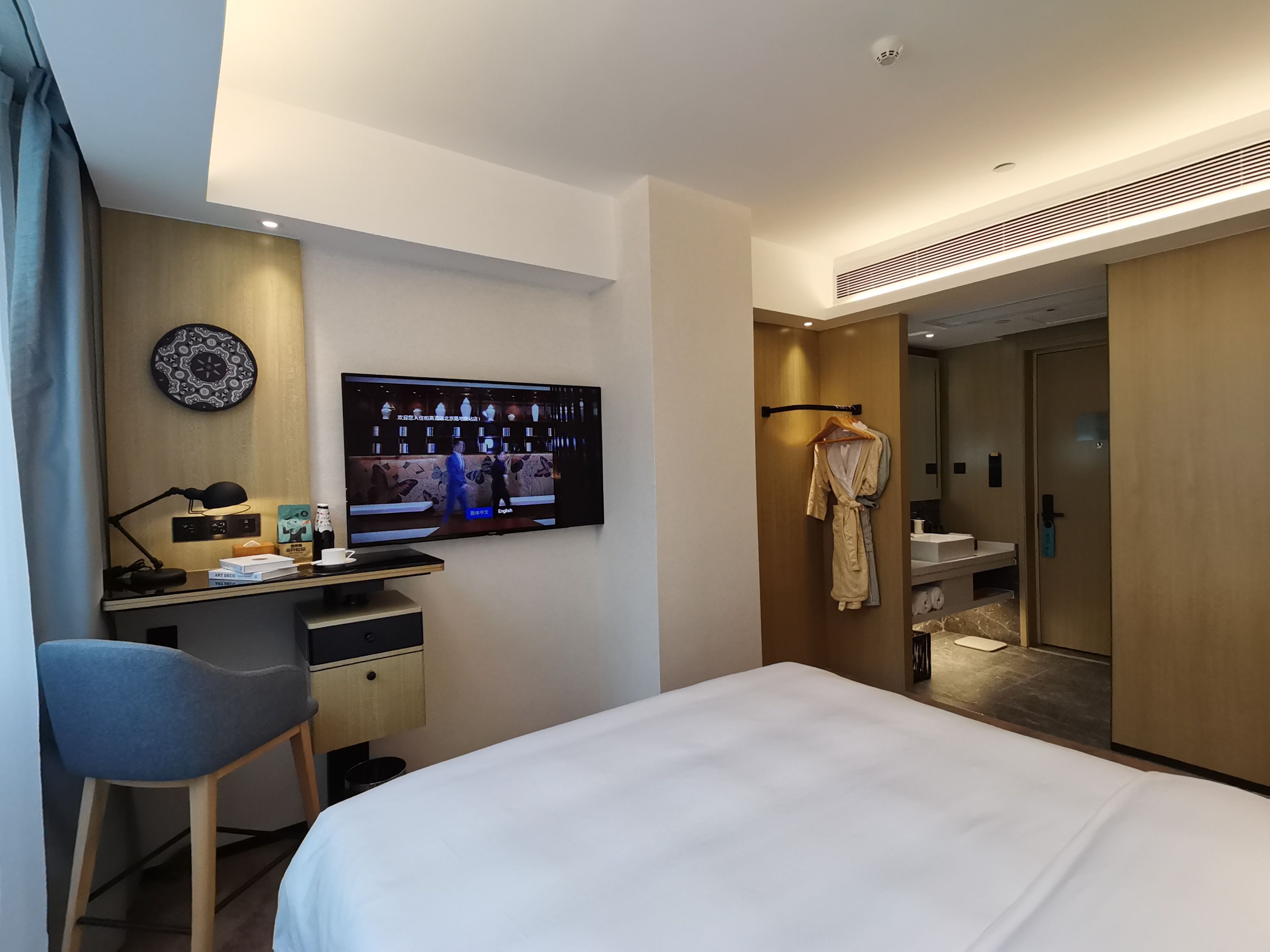 A hotel room with a bed, a desk with a computer, a TV, and a bathroom with a robe hanging.